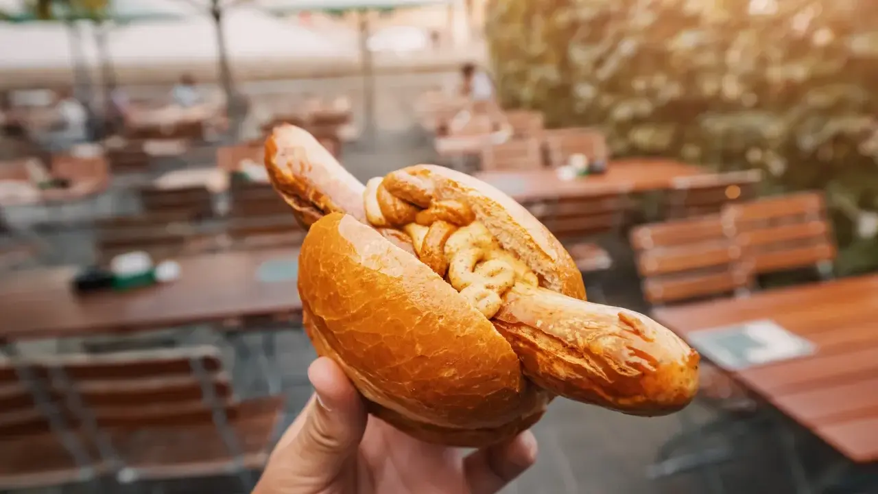 Eine Hand hält eine Bratwurst im Brötchen mit Senf. Ein leckerer Snack, aber die Bratwurst Kalorien sollte man im Auge behalten.