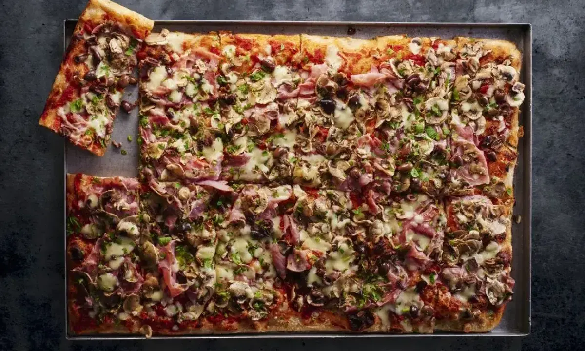 Prostokątna pizza 4 sery z pieczarkami, szynką i oliwkami, pokrojona na kwadraty, z jednym kawałkiem odłożonym na bok.