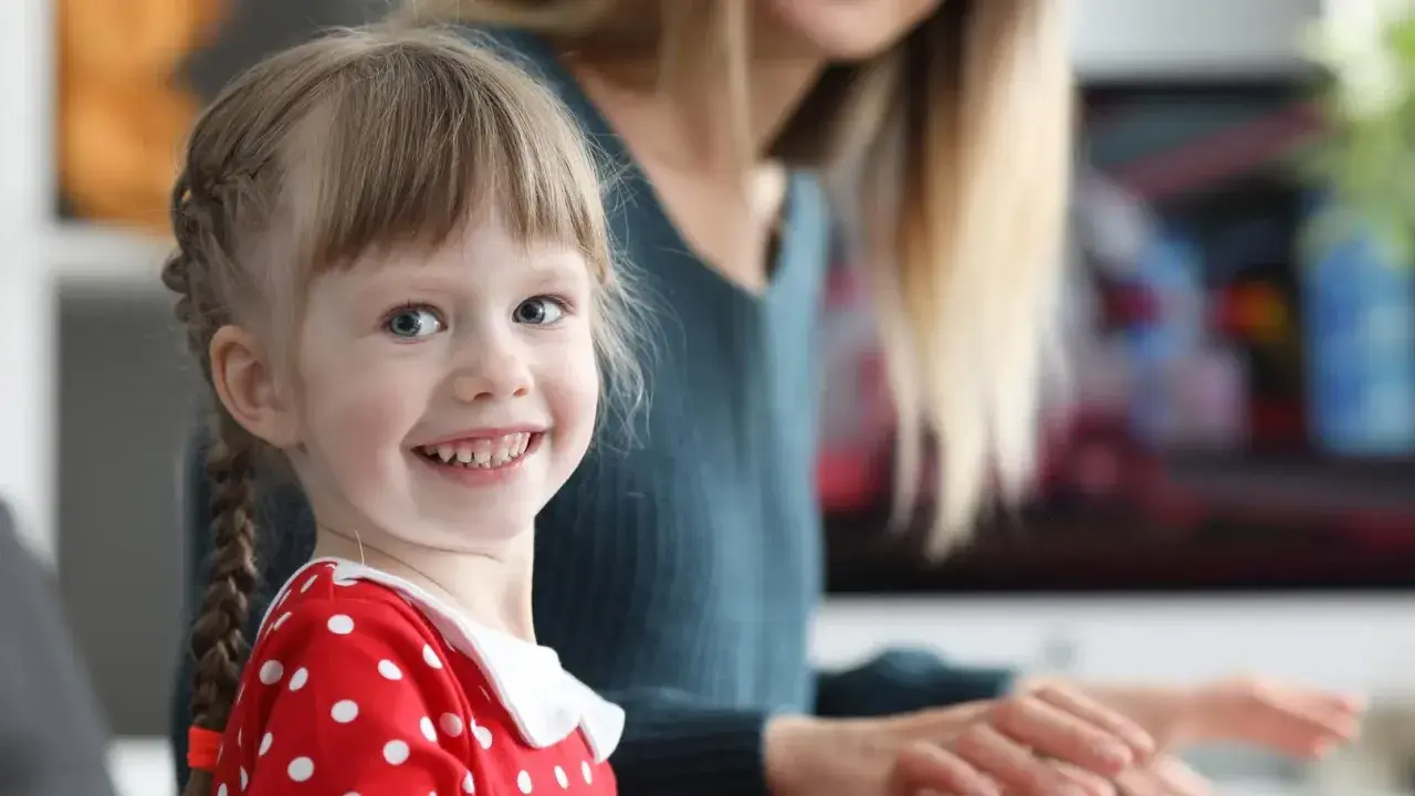 Uśmiechnięta dziewczynka w czerwonej sukience w groszki, z warkoczykami, uczy się kindersztuby od mamy.