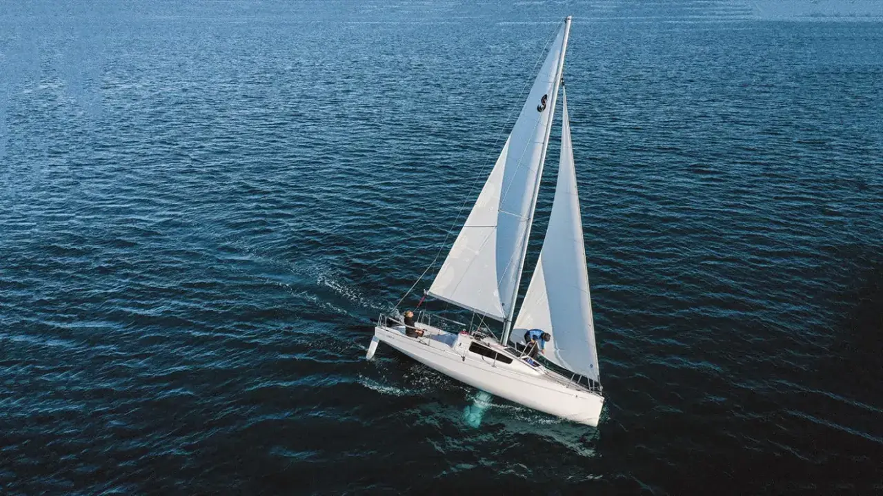 Un voilier quille relevable navigue sur une mer calme, ses voiles blanches gonflées par le vent. Deux personnes sont à bord.