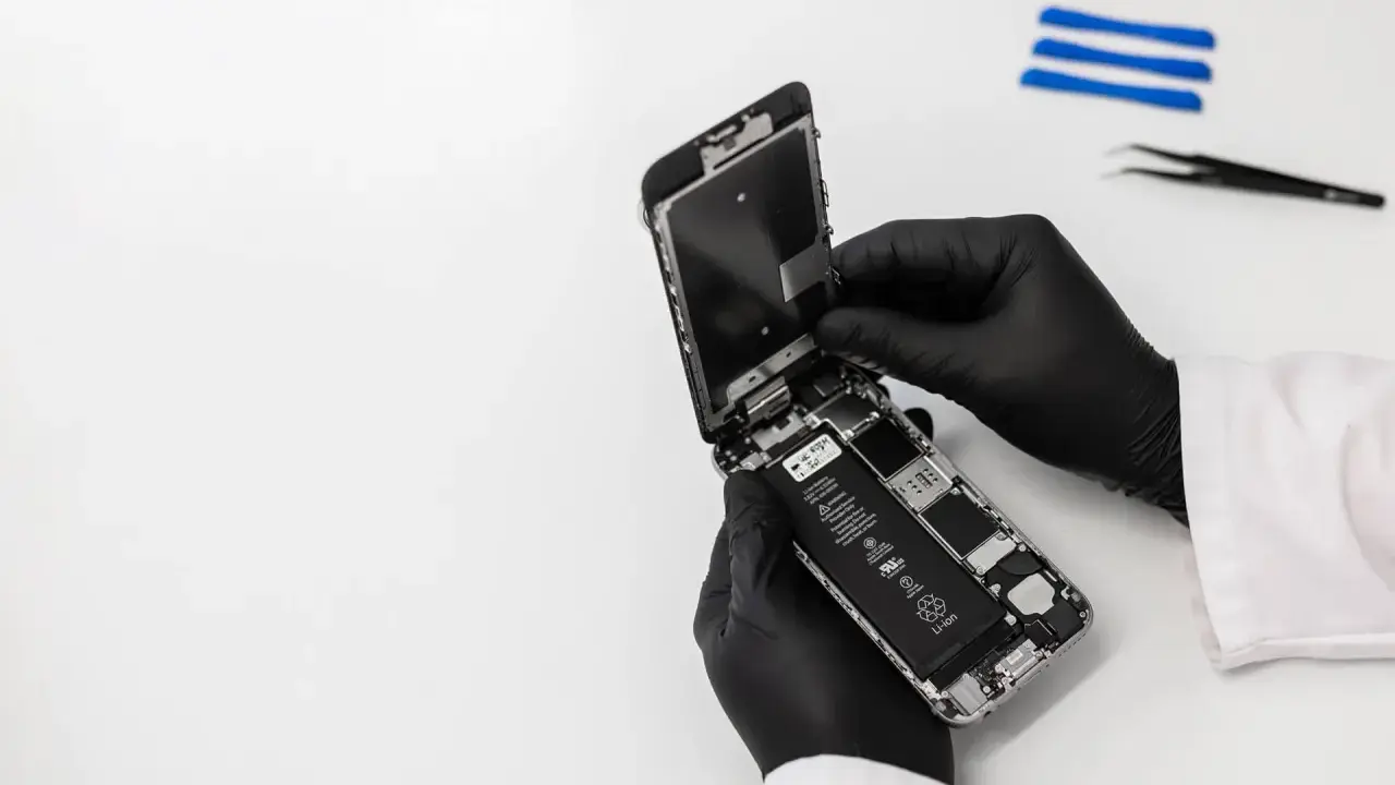 Ręce w czarnych rękawiczkach rozbierają iPhone'a, który wpadł do wody, ale działa. Widać baterię i wnętrze telefonu.
