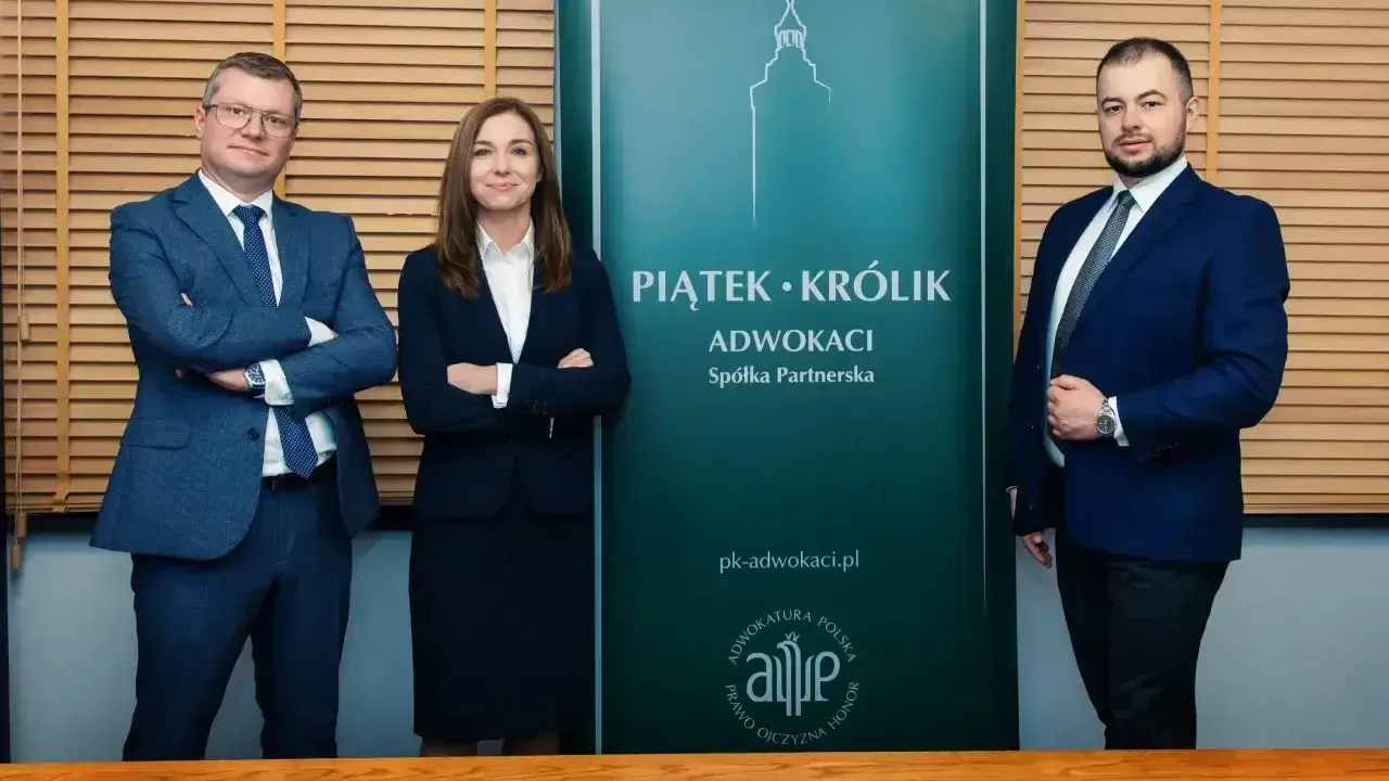 Trzy osoby w garniturach stoją przed zielonym banerem kancelarii "Piątek • Królik Adwokaci".