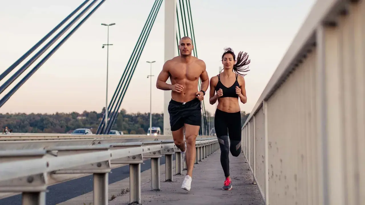 Ein Mann und eine Frau joggen auf einer Brücke. Beide haben sichtbare bauchmuskeln, ein Zeichen für ihr intensives Training.