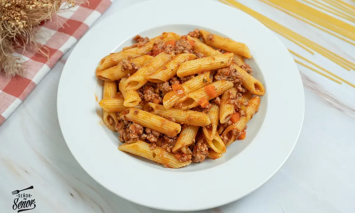 Plato de macarrones con carne picada y salsa de tomate, decorado con un paño a cuadros y espaguetis secos al fondo.