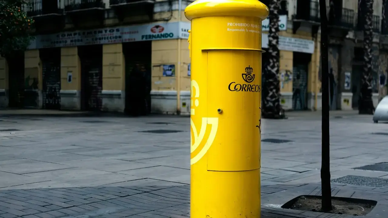 Jasnożółta skrzynka pocztowa Correos, symbol poczty Hiszpanii, stoi na brukowanym placu przed budynkami.