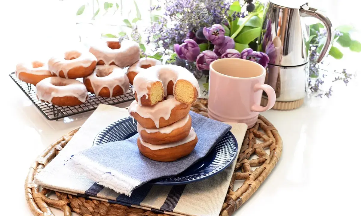 Deliciosos donuts caseros glaseados, apilados en un plato con una taza rosa y flores moradas.