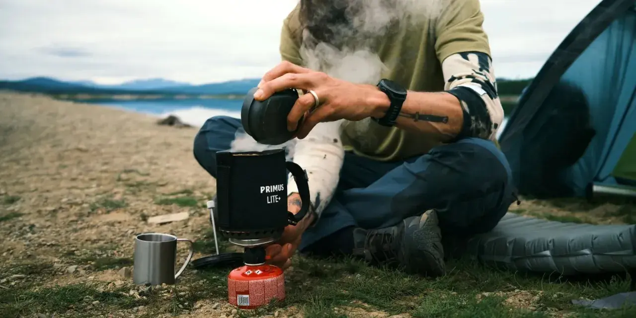 En optimus hiker njuter av en varm dryck vid en sjö. Ånga stiger från koppen medan personen sitter bredvid ett tält.