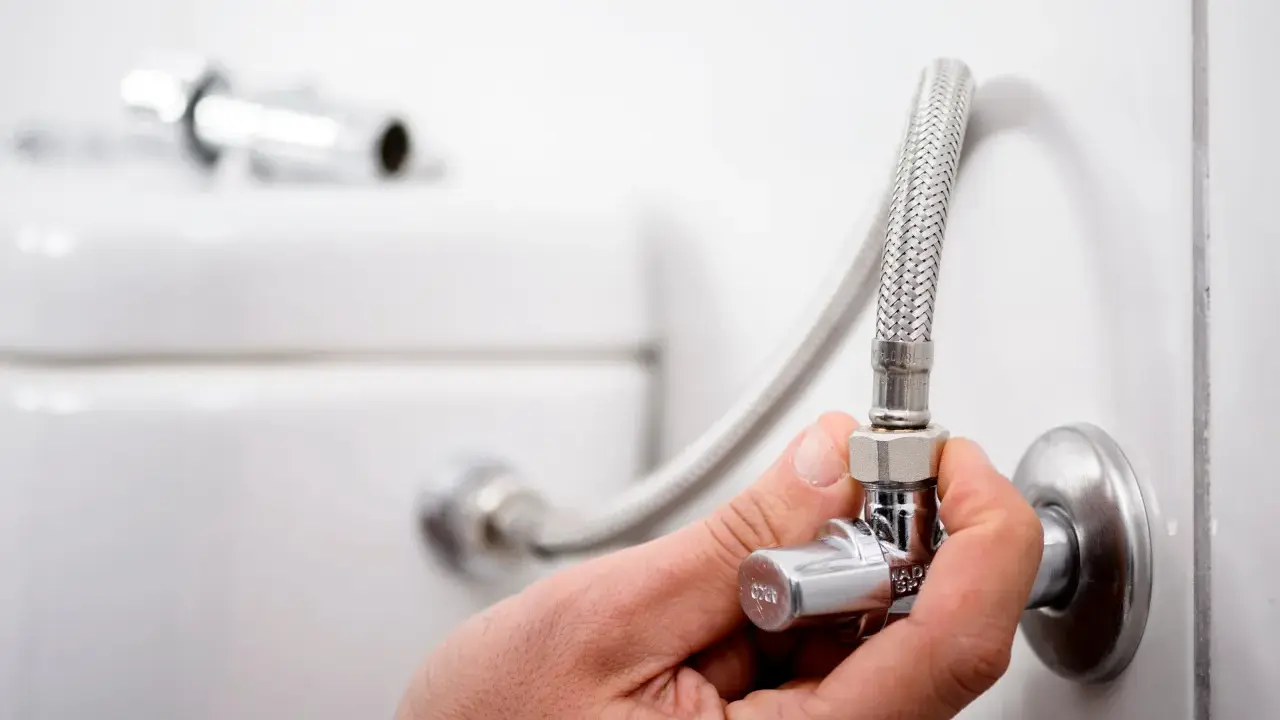 Mano ajustando la llave de paso del agua, que es comunitaria, conectada a una manguera flexible y al inodoro.