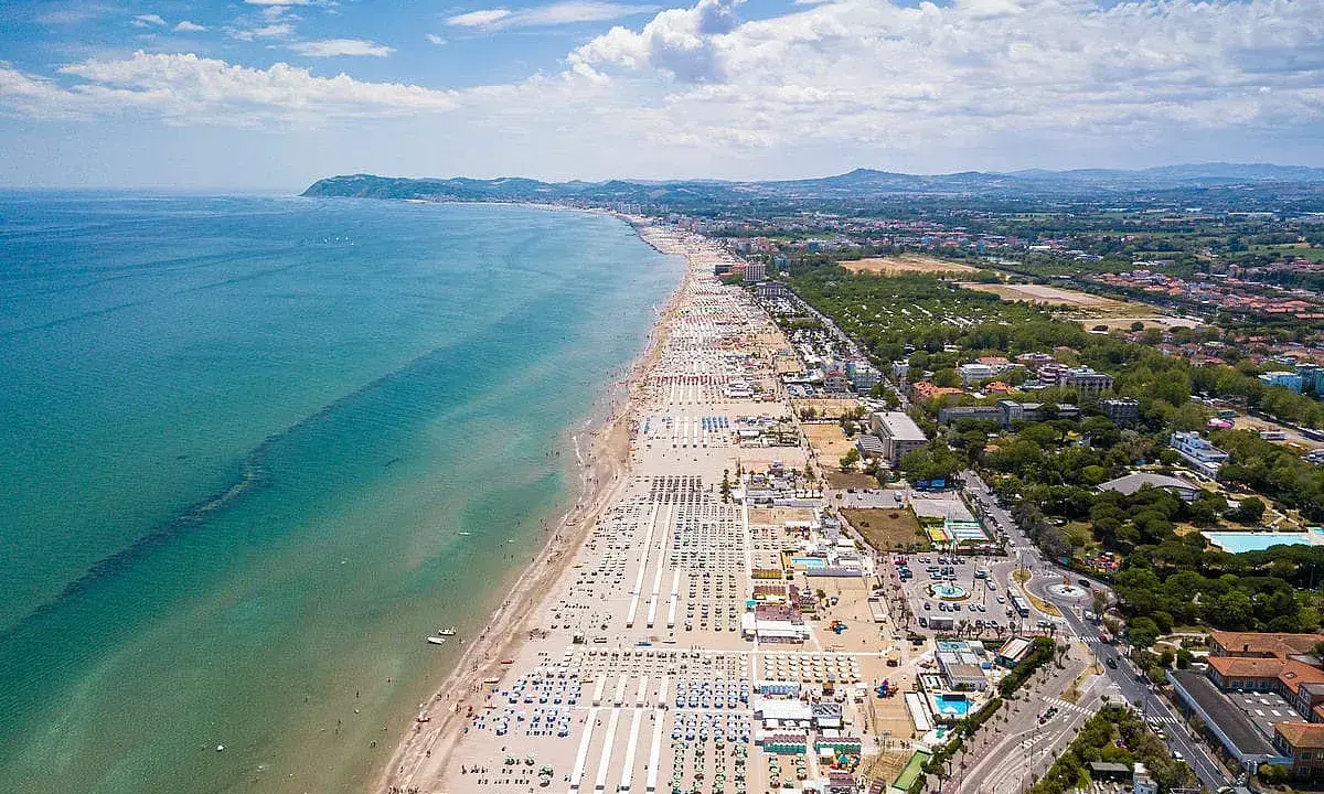 Vista aerea della Riviera Romagnola con spiaggia affollata, mare azzurro e verdeggiante entroterra.