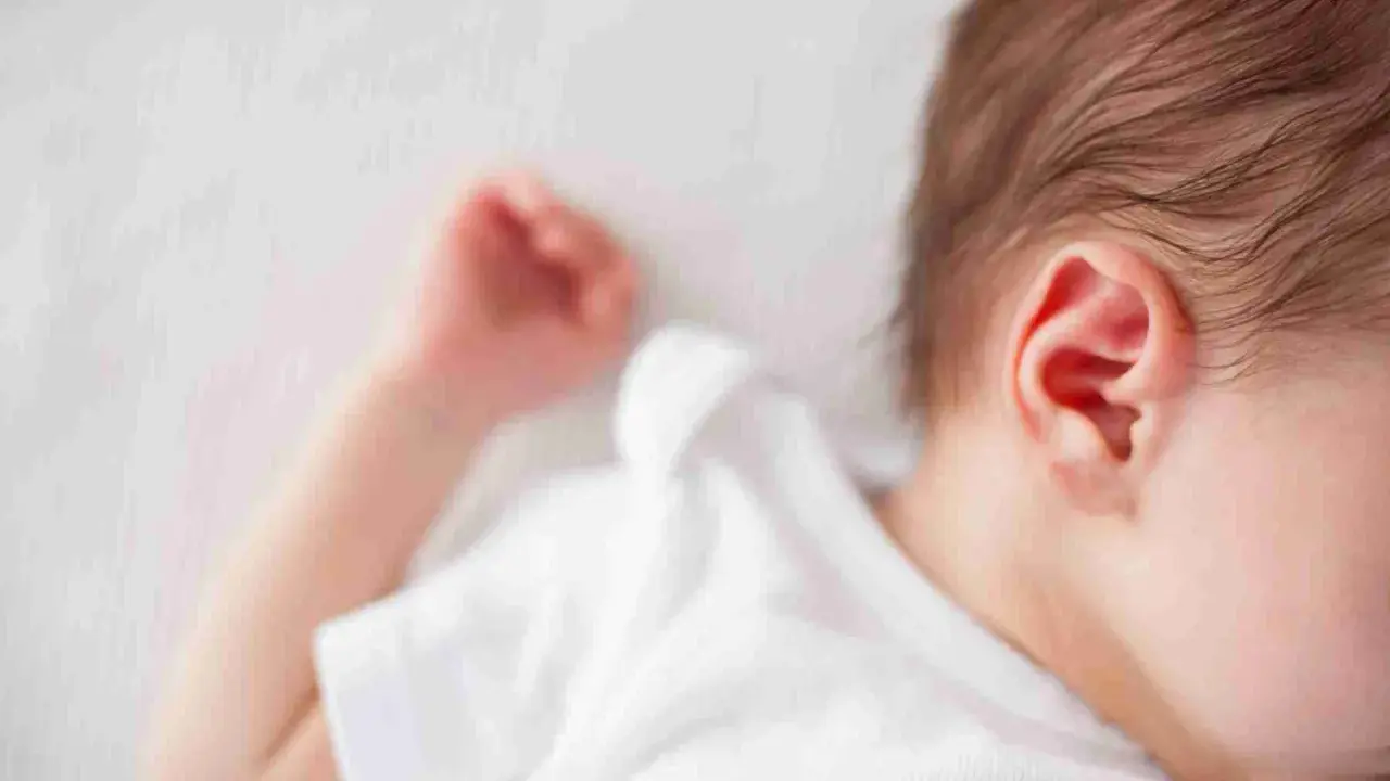 Un neonato dorme troppo, con la testolina appoggiata su un morbido lettino bianco. Si intravede un orecchio rosa e una manina.
