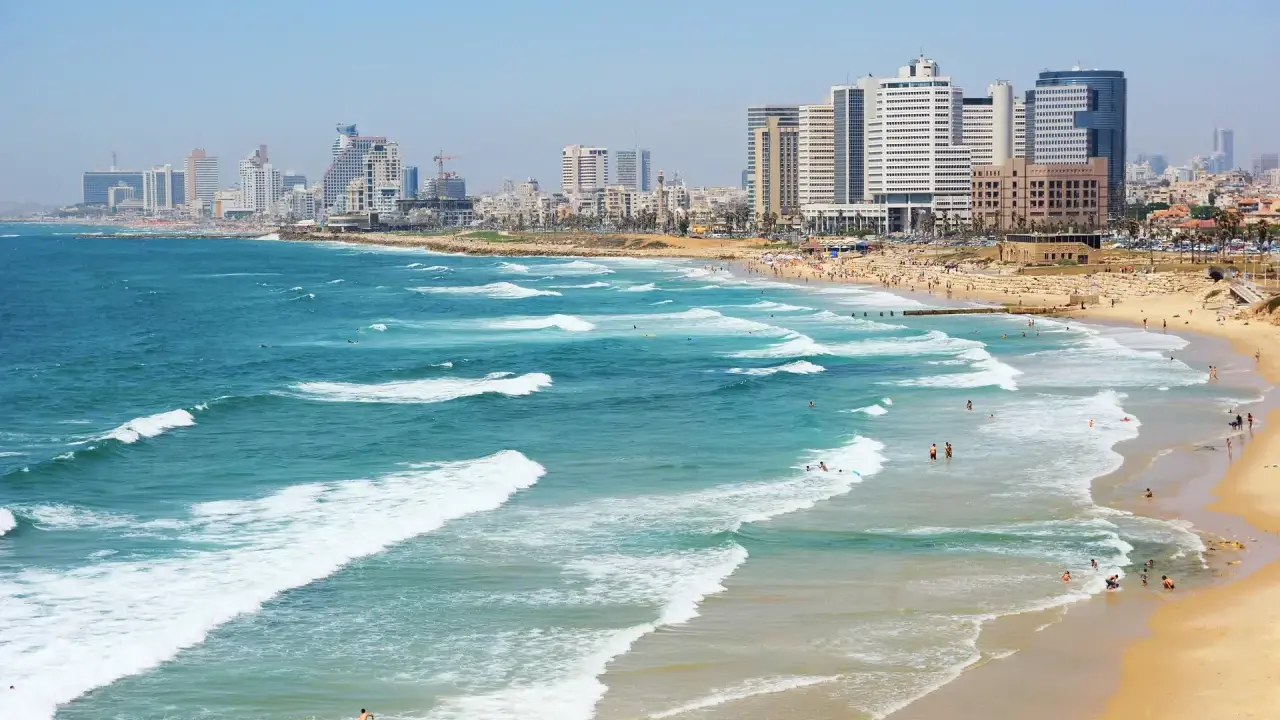 Plaża w Tel Awiwie, z falami Morza Śródziemnego i nowoczesnym miastem w tle. Ludzie kąpią się i opalają, co może sugerować, że **czy w Izraelu jest bezpiecznie** dla turystów.