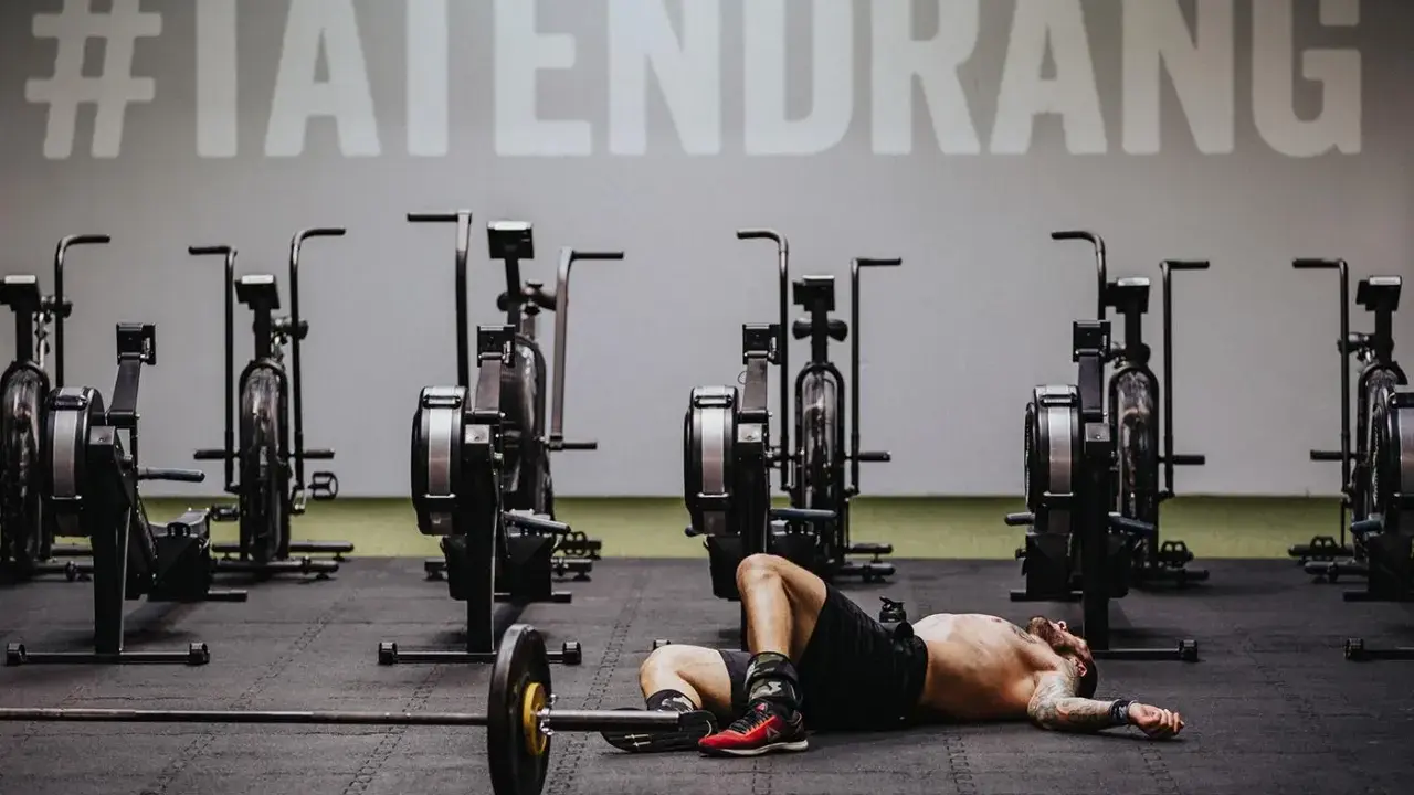 Mann liegt erschöpft auf dem Boden neben einer Hantelstange, umgeben von Trainingsgeräten. Ein Zeichen an der Wand verkündet "#TATENDRANG" – ein Symbol für den Wiedereinstieg in den **Sport nach Corona**.