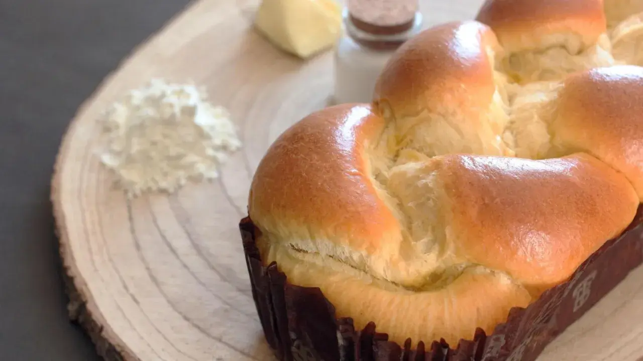 Un pan brioche dorado y esponjoso, listo para ser disfrutado. A su lado, ingredientes para su elaboración: harina, mantequilla y sal.