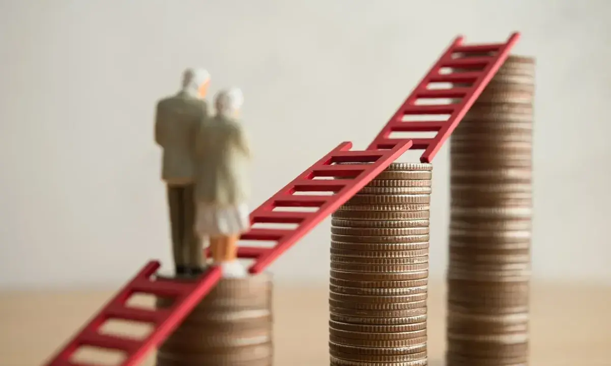 Figurki starszej pary stoją na stosach monet, połączonych czerwoną drabiną. Symbolizuje to nowe zasady przejścia na emeryturę i drogę do finansowej stabilności.