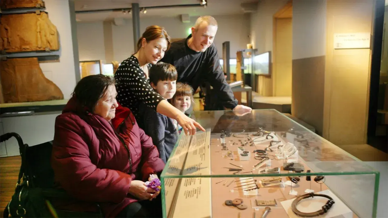 A family, including a woman in a wheelchair, examines artifacts in a museum display, perhaps inspired by famous art curators.
