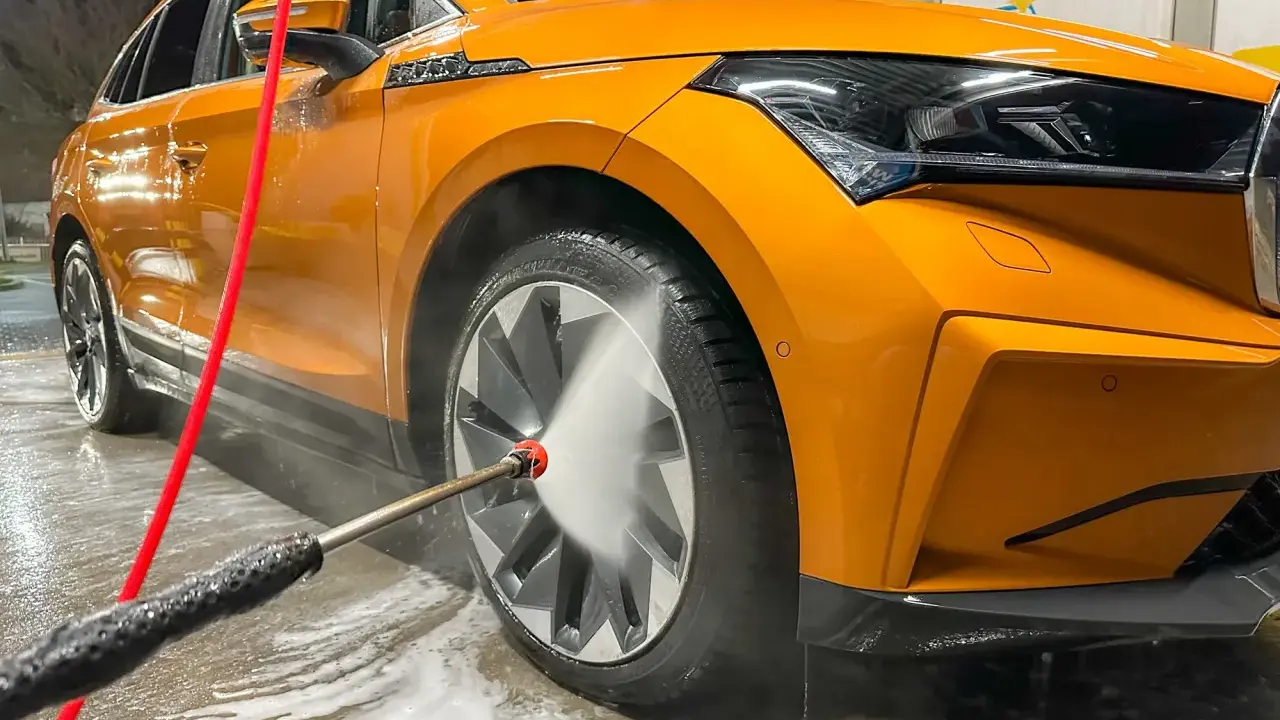 Pomarańczowe auto w trakcie mycia. Woda pod ciśnieniem spływa po karoserii i felgach, tworząc pianę.