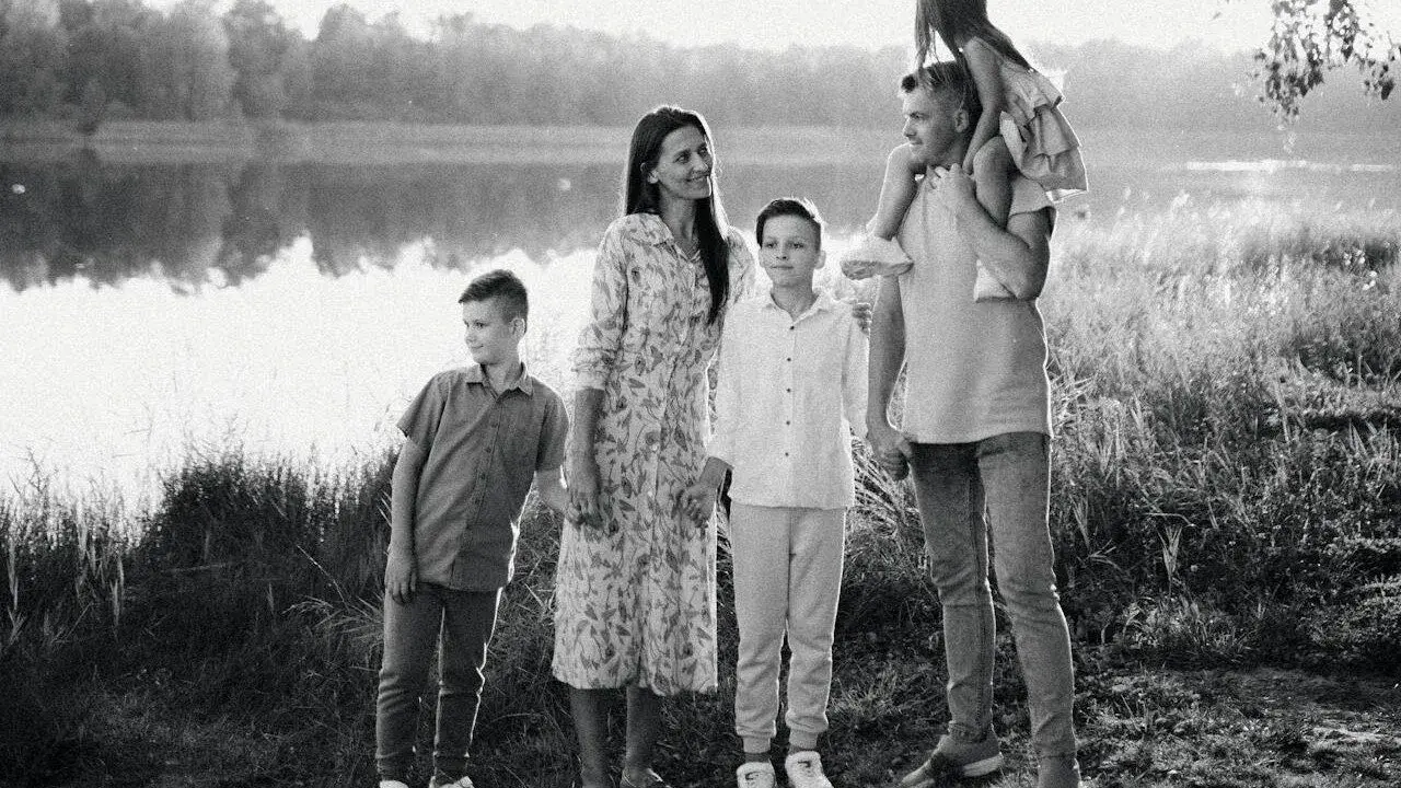 Czarno białe zdjęcie. Rodzina na tle jeziora. Ojciec niesie córkę na barana, obok mama i dwaj synowie.