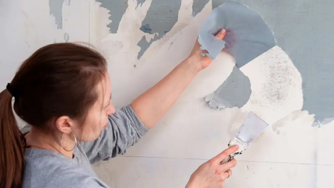 Une femme utilise une spatule pour décoller du papier peint ancien.