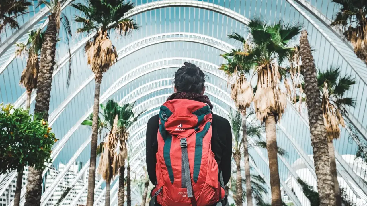 Viaggiatore con zaino rosso sotto palme e architettura moderna. La traduzione di "daffodils" è narcisi, ma qui non ci sono fiori.