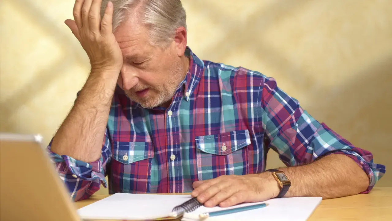 Uomo anziano con mano sulla fronte, frustrato dal non ricordare le cose appena fatte, con quaderno e penna.