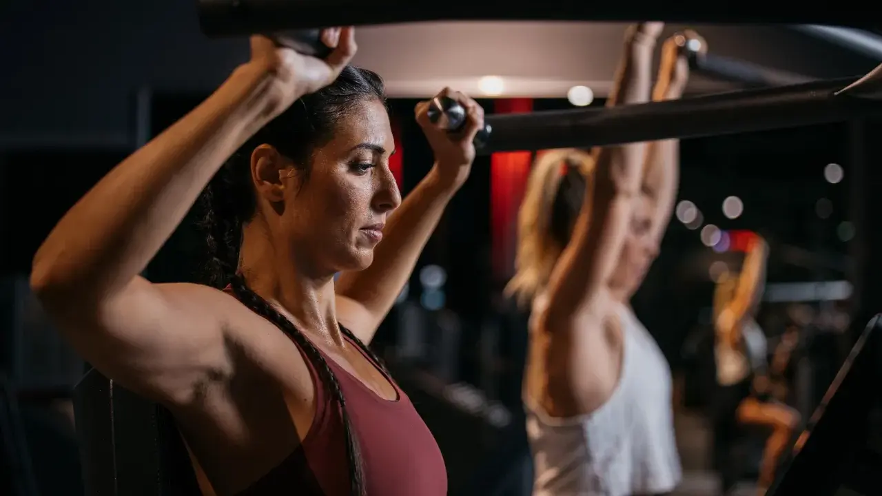 Zwei Frauen trainieren im Fitnessstudio. Sie nutzen eine Maschine, die zeigt, welche Muskelgruppen zusammen trainieren.