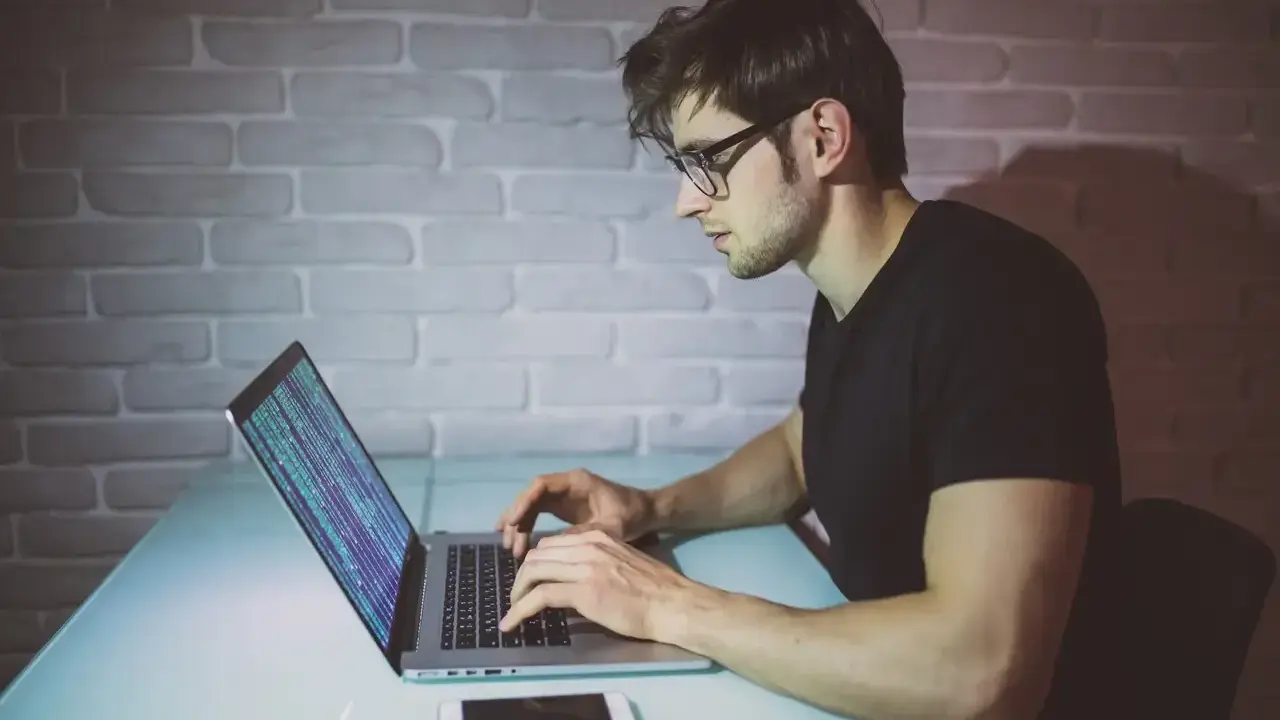 Młody mężczyzna w okularach pisze kod na laptopie. To jego pierwszy krok, by dowiedzieć się, jak zostać informatykiem.