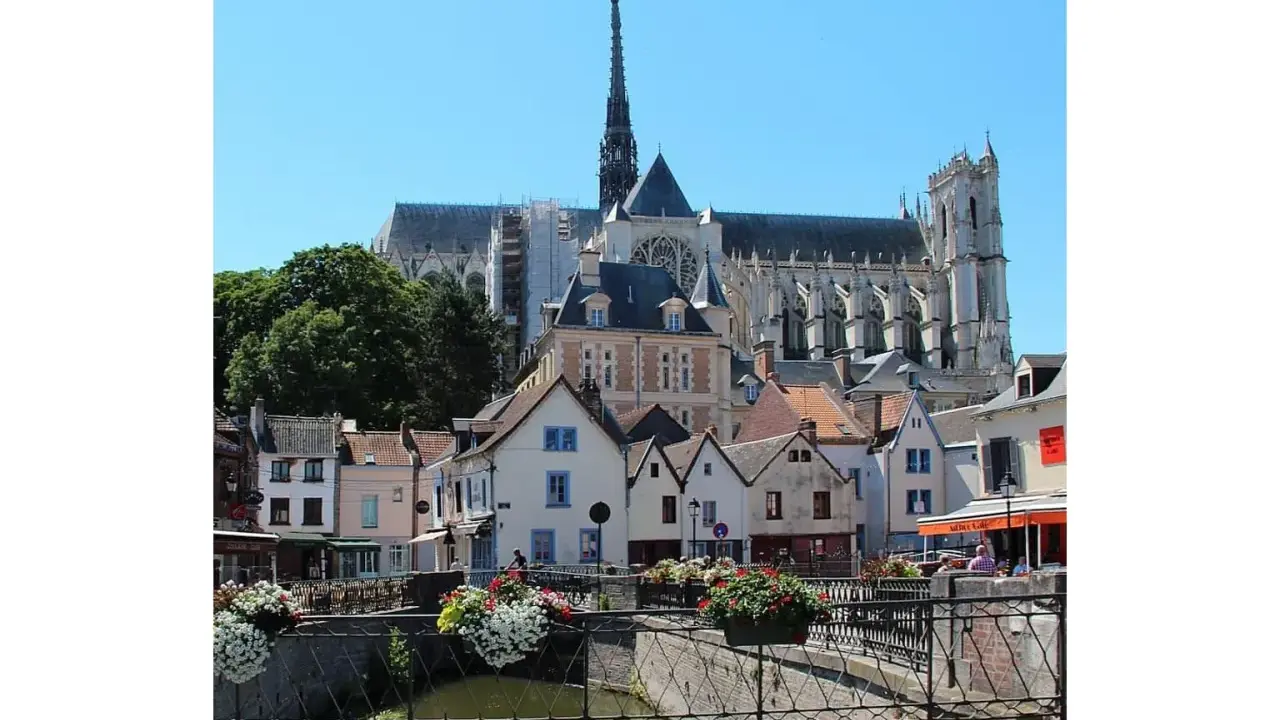 Blick auf malerische Häuser und die Kathedrale in Amiens, Frankreich. Blumenkästen schmücken eine Brücke über einen Kanal.