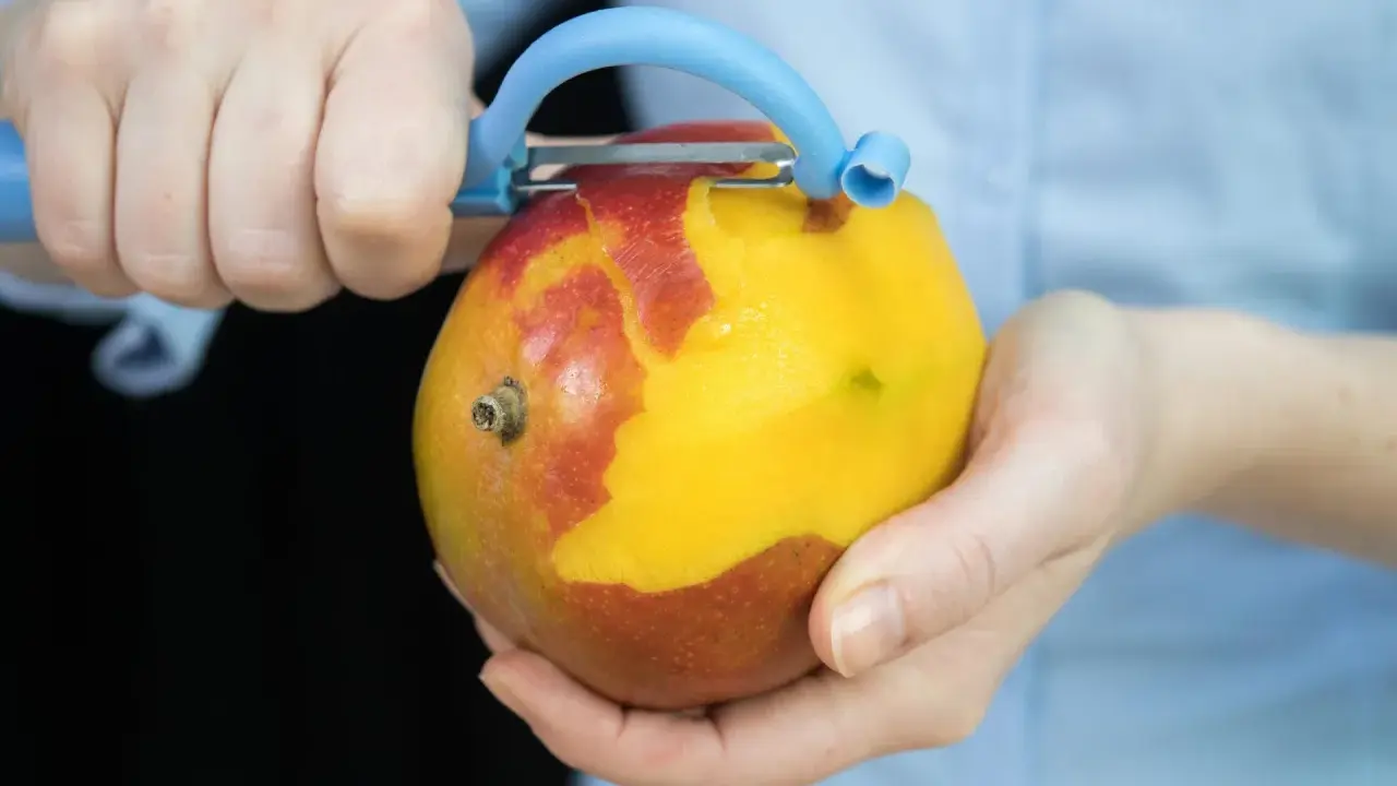 Hände schälen eine reife Mango mit einem blauen Sparschäler. Das Fruchtfleisch leuchtet gelb, während die Schale rot und gelb ist.