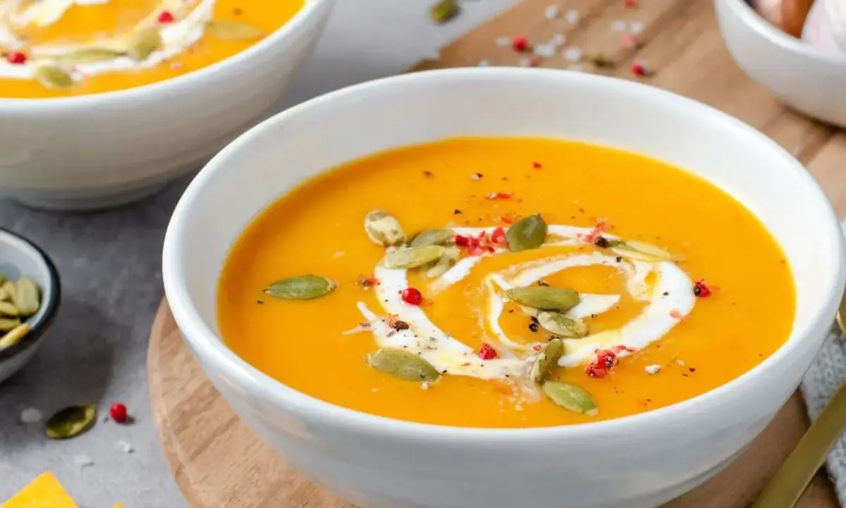 Velouté de courge butternut orange servi dans un bol blanc, garni de crème, de graines de courge et de poivre rose.