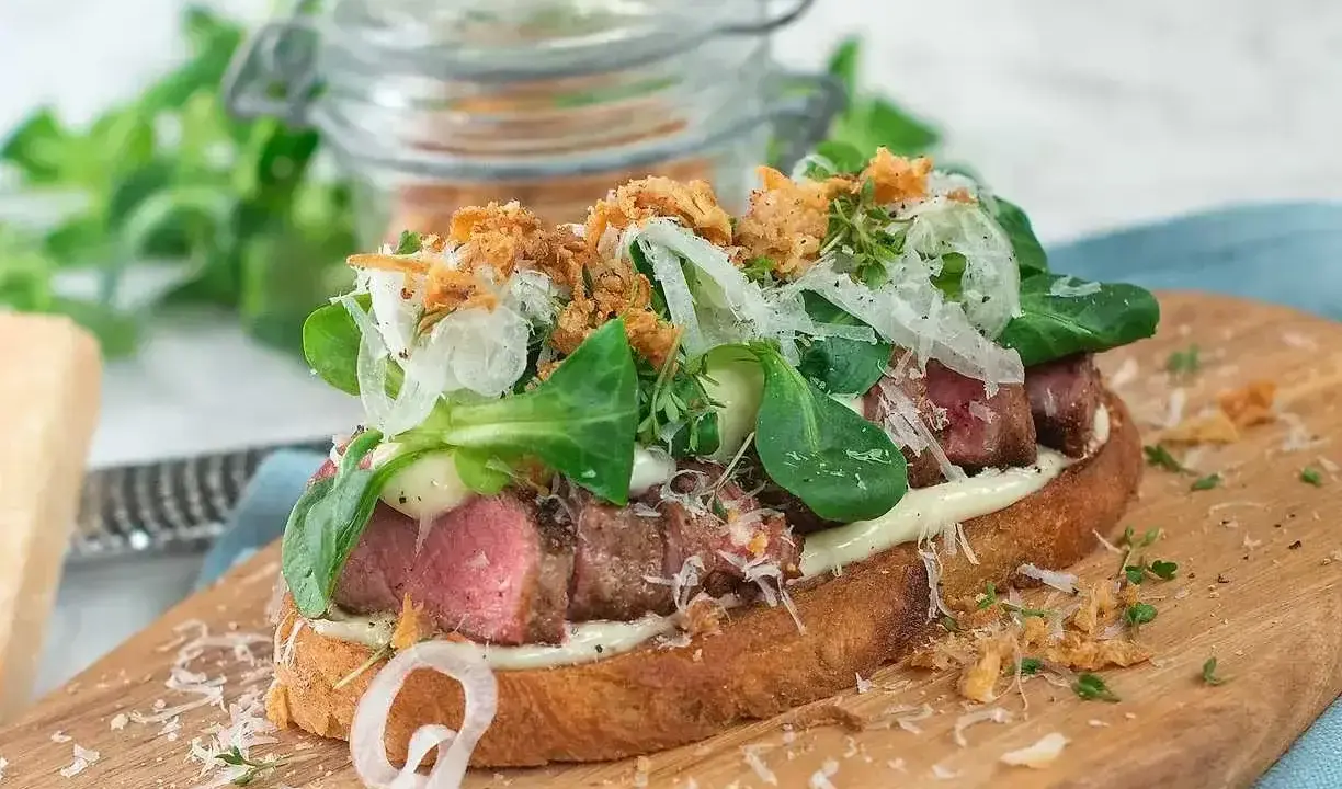 En saftig steak sandwich med möra skivor av biff, krämig majonnäs, färska gröna blad, krispig lök och riven parmesanost på rostat bröd.