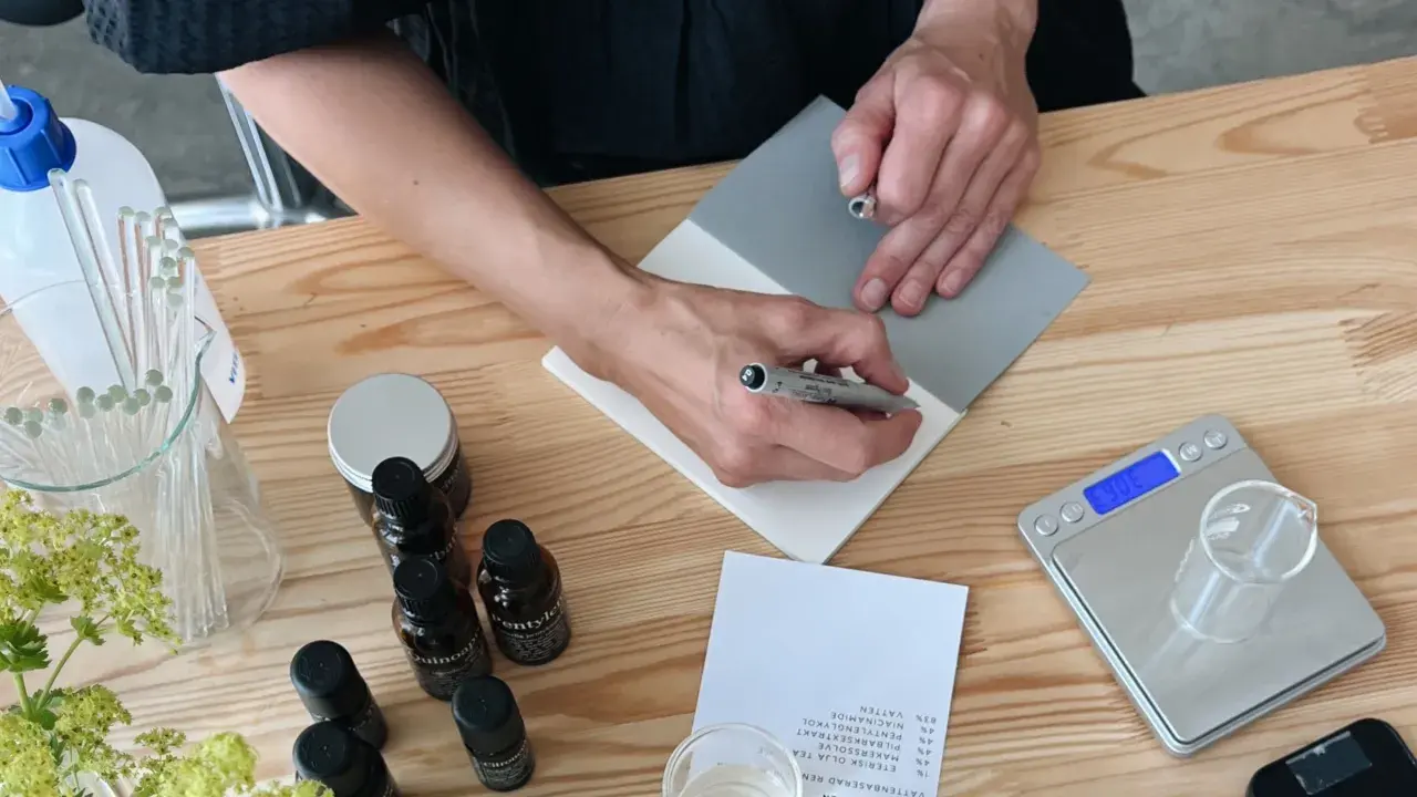 Person skriver recept för ekologiska doftpinnar. Ingredienser och verktyg ligger på ett träbord.