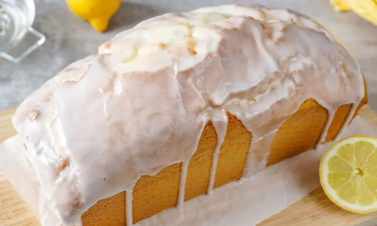 Ein saftiger Zitronenkuchen in Kastenform, überzogen mit süßem Zuckerguss. Ein Zitronenviertel liegt daneben.