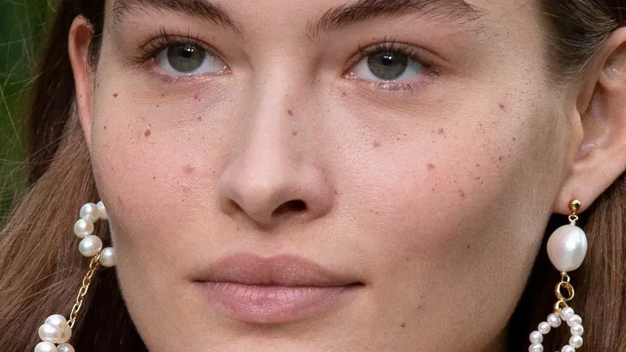 Primer plano de un rostro femenino con pecas naturales salpicando su piel. Lleva pendientes de perlas colgantes.