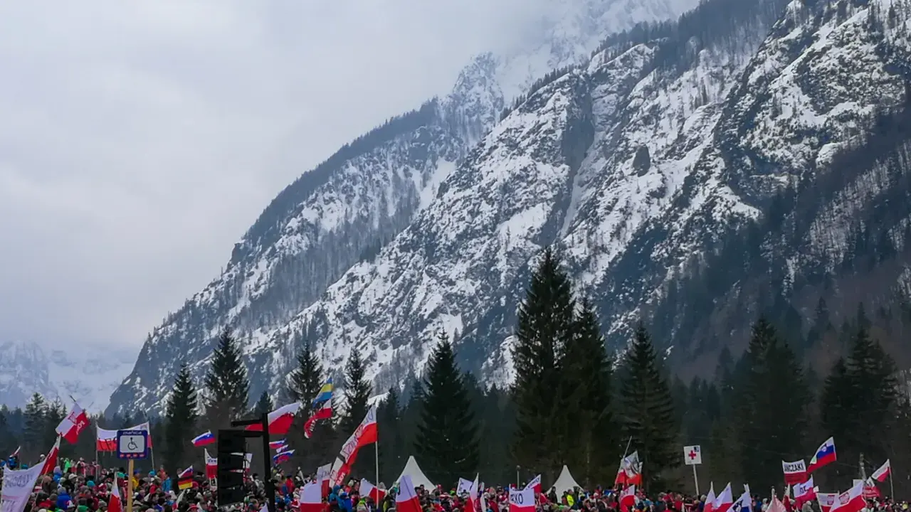 Tłum kibiców z flagami na tle ośnieżonej góry, atmosfera na skoczni w Planicy.