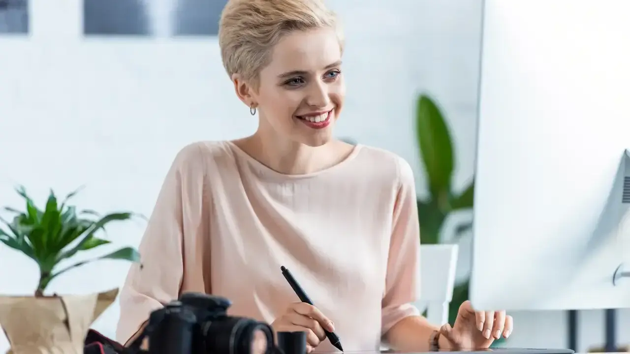 Uśmiechnięta kobieta z krótkimi blond włosami pracuje przy komputerze, obok aparat fotograficzny. Sprawdza najnowsze oferty pracy w Pile.