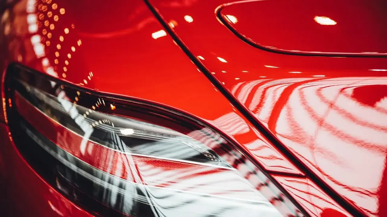 Błyszczący, czerwony lakier samochodu, odbijający światła. Widać fragment lampy. To praca detailera, który doprowadził auto do perfekcji.