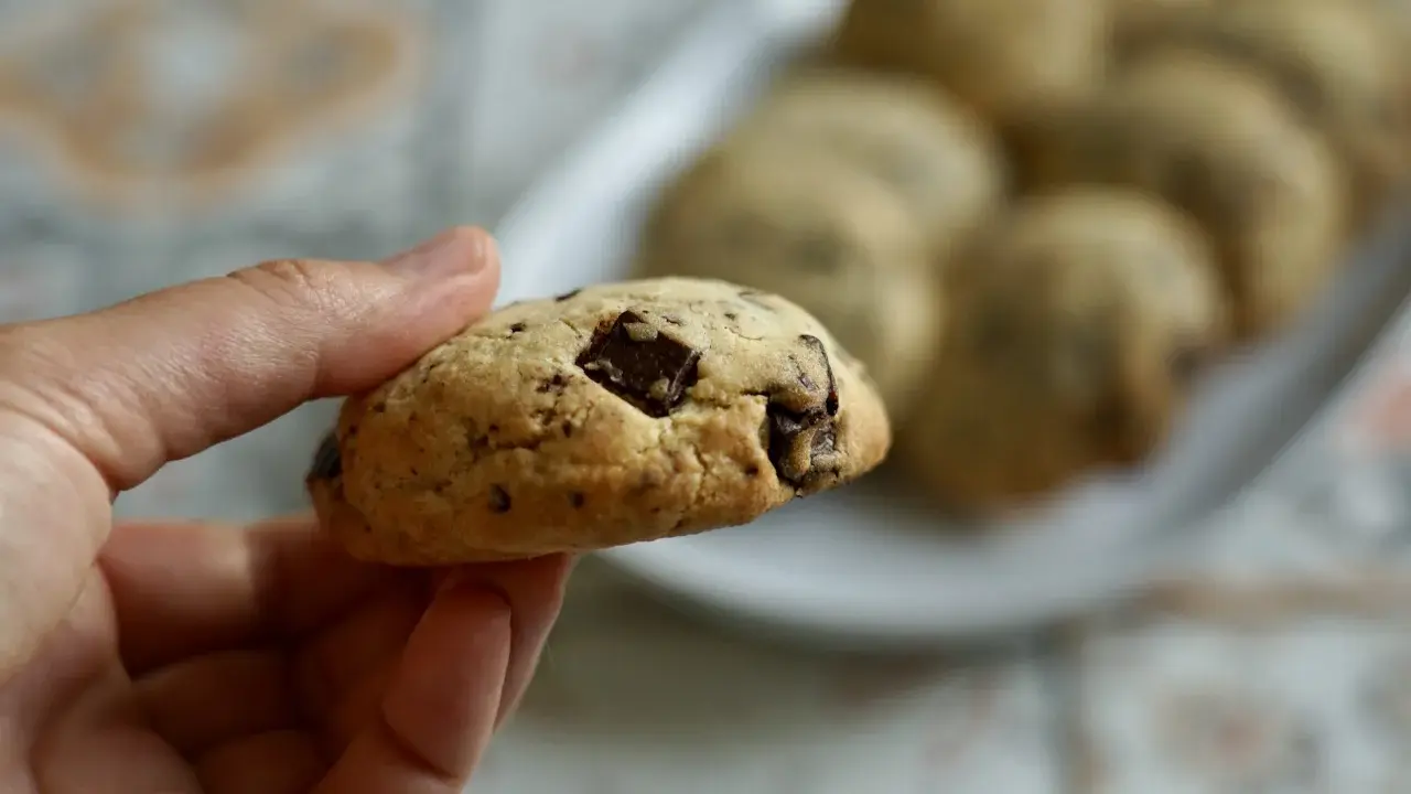 Une main tient un cookie épais et moelleux aux pépites de chocolat. D'autres cookies sont disposés sur une assiette en arrière-plan.