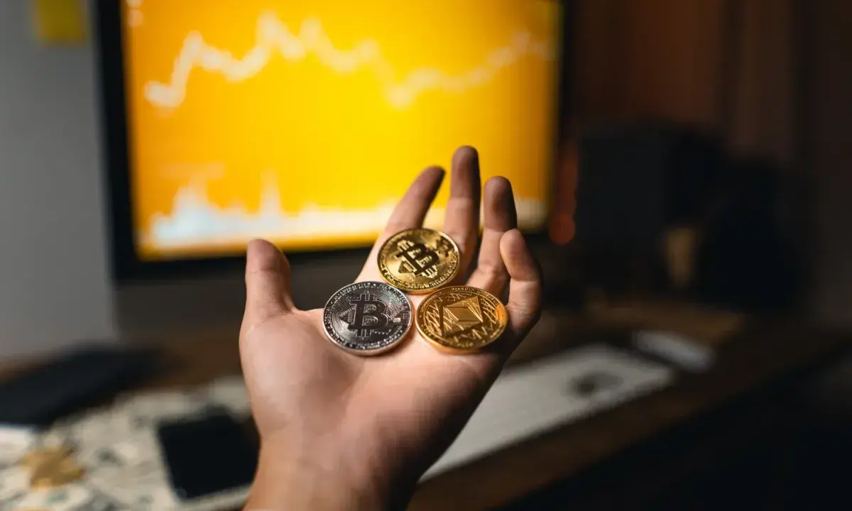Dłoń trzyma trzy monety kryptowalut: Bitcoin, Ethereum i inną. W tle wykres na monitorze, sugerujący handel na kantory kryptowalut.