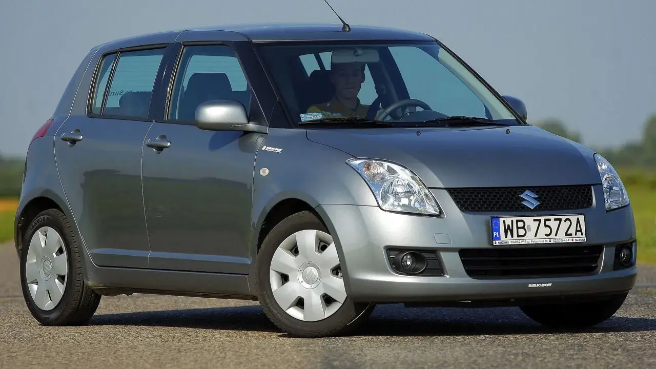 Szary Suzuki Swift 2007 z kierowcą za kierownicą, zaparkowany na drodze.