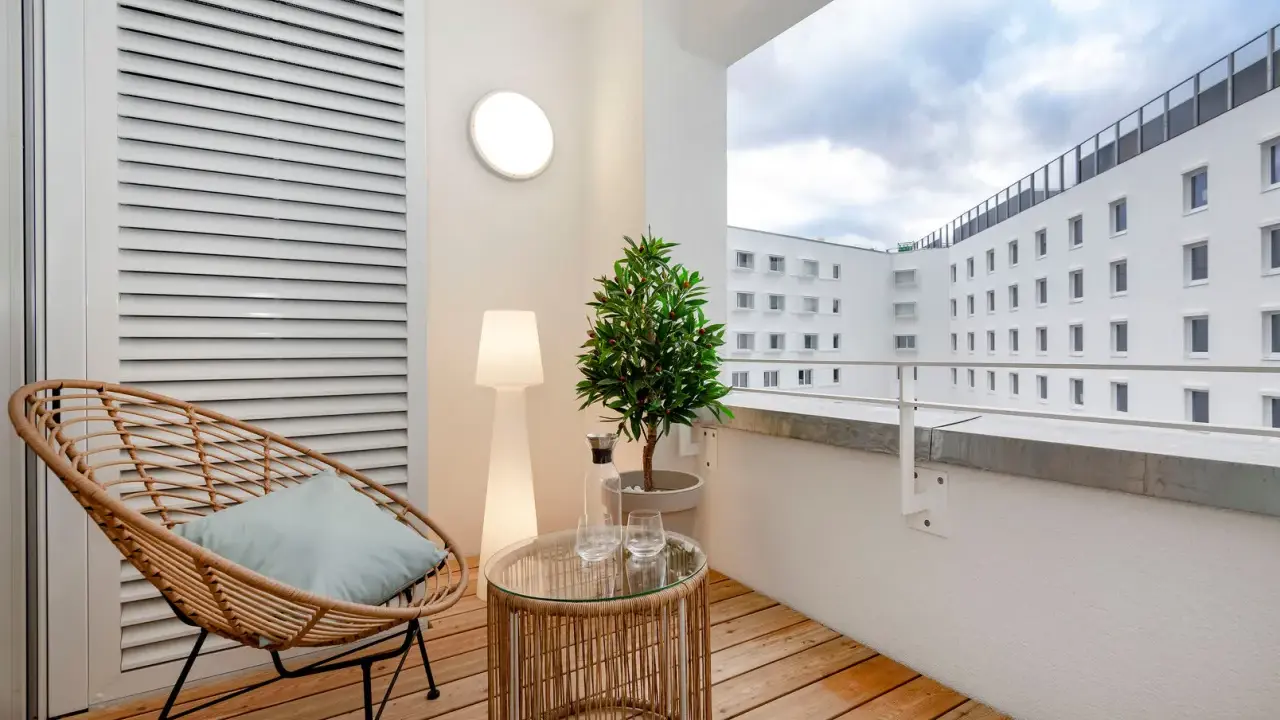 Przytulny balkon z drewna, z wiklinowym fotelem, stolikiem i rośliną. W tle białe budynki.