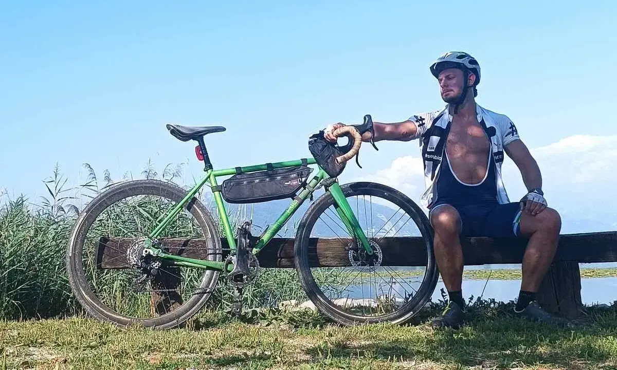Ciclista riposa accanto alla sua bici verde, pensando alle tabelle per dimagrire in bicicletta. Cielo azzurro e lago sullo sfondo.
