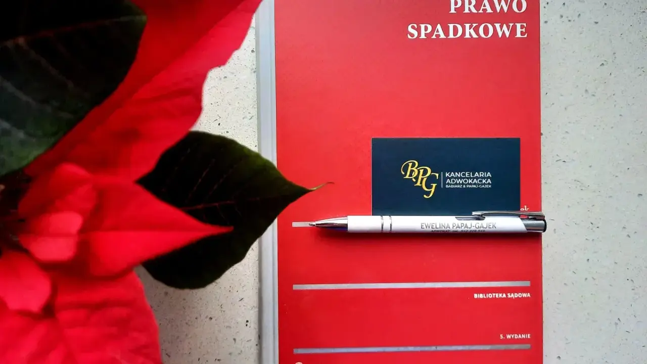 Czerwona książka "Prawo spadkowe" obok poinsecji. Długopis z nazwiskiem adwokatki i wizytówka kancelarii sugerują tematykę prawną, być może związaną z zapisem windykacyjnym.