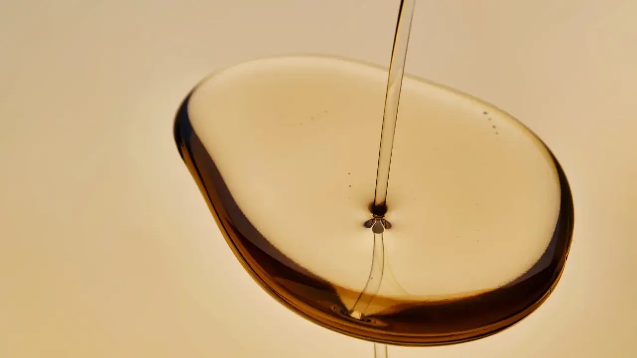 Un filet d'huile de karanja, d'une couleur ambrée, tombe sur une surface liquide, créant des reflets sombres. Attention, l'huile de karanja peut présenter un danger.