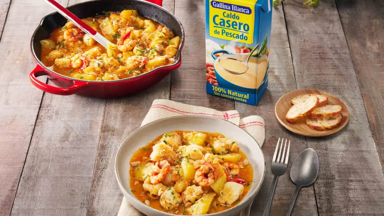 Rape a la marinera con patatas y gambas, servido en cazuela roja y plato. Al lado, caldo de pescado Gallina Blanca y pan tostado.