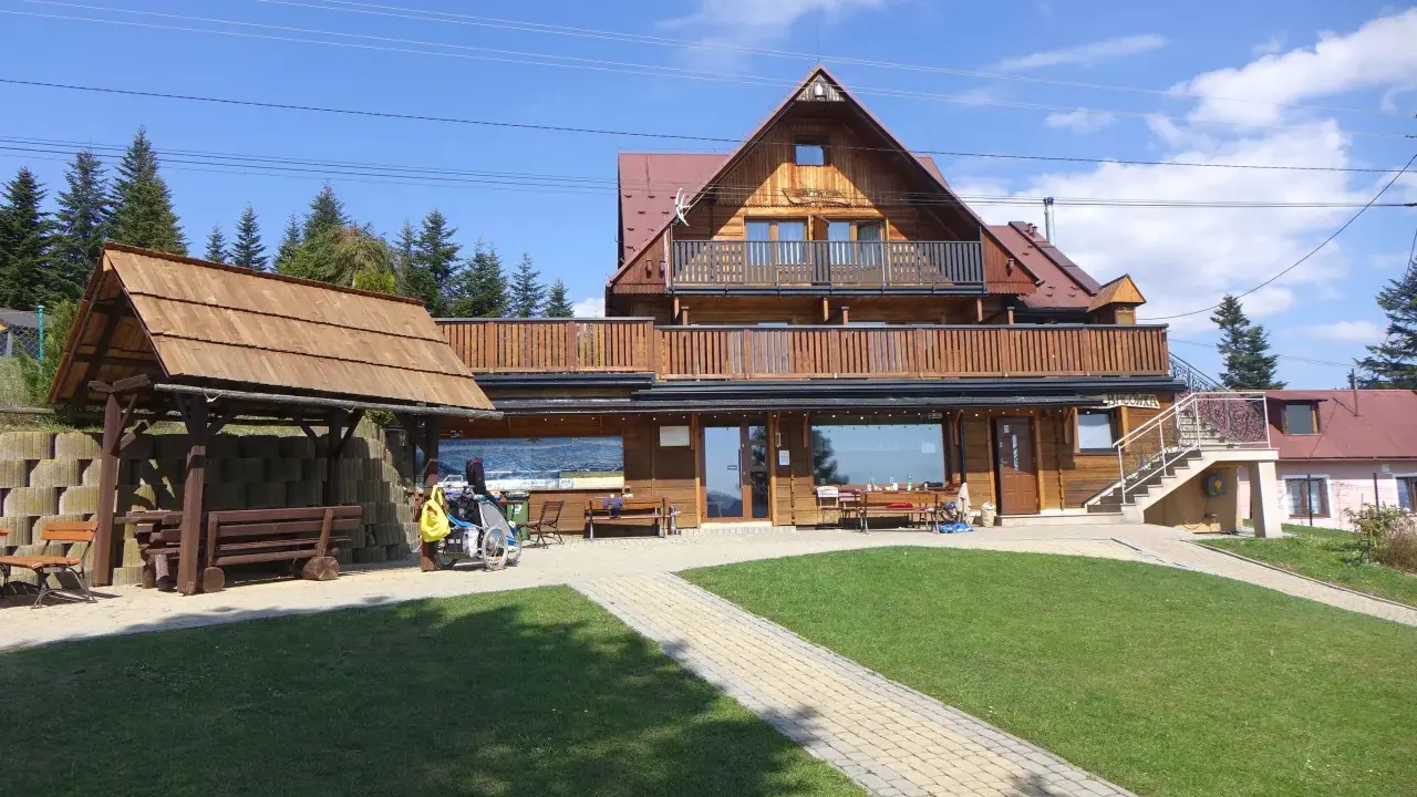 Drewniana bacówka na obidzy z balkonem i tarasem, otoczona zielenią i ścieżkami. Obok wózek inwalidzki z żółtą torbą.