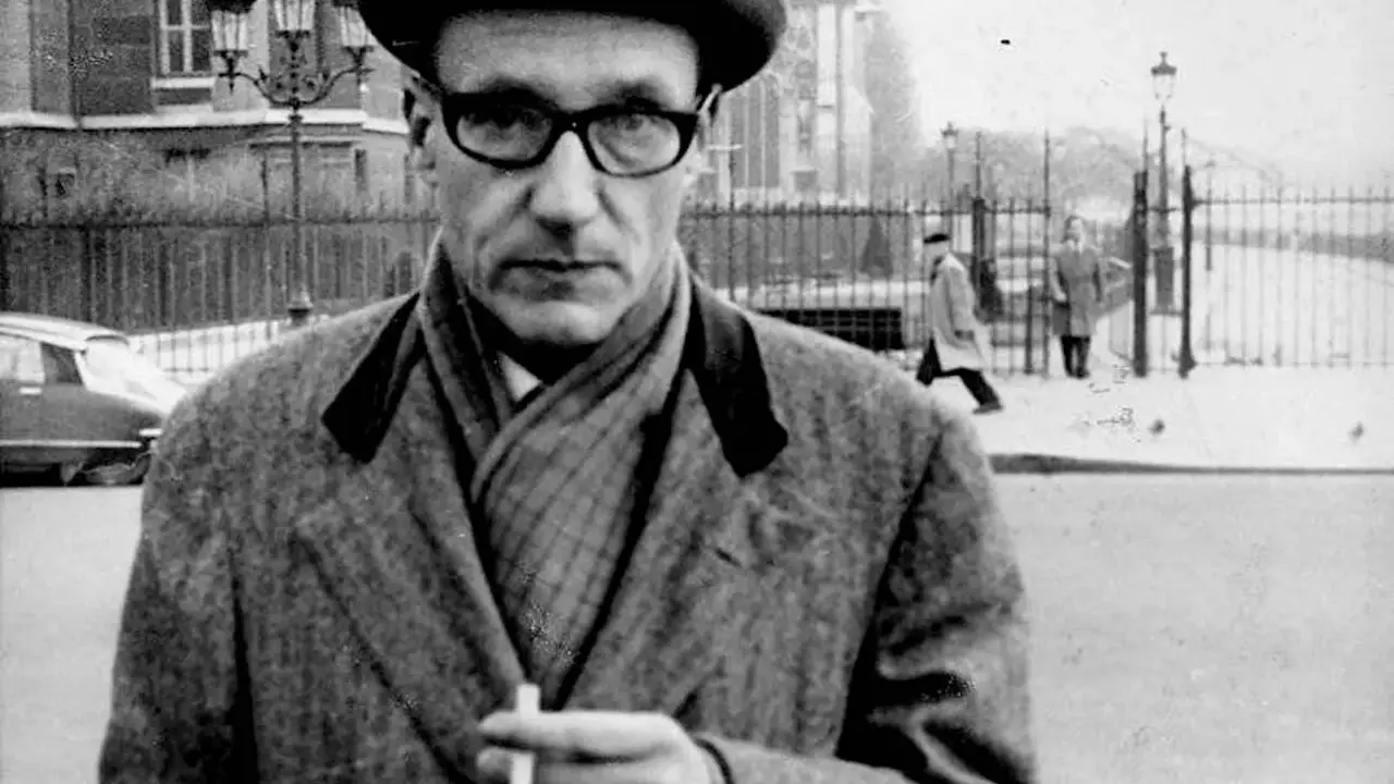 William Burroughs, con cappello e occhiali, tiene una sigaretta in mano, passeggiando in una strada urbana con persone sullo sfondo.