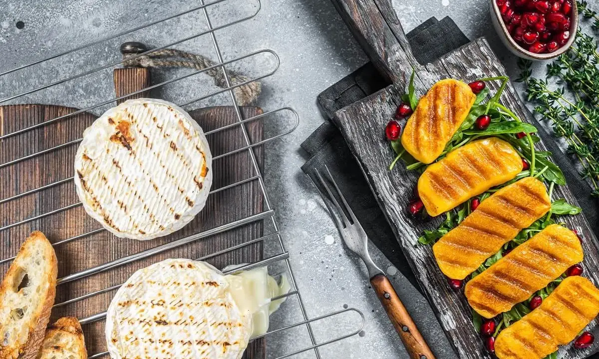 Dwa okrągłe sery camembert grillowane na ruszcie, obok pieczywo i grillowane kawałki sera na desce z granatem.