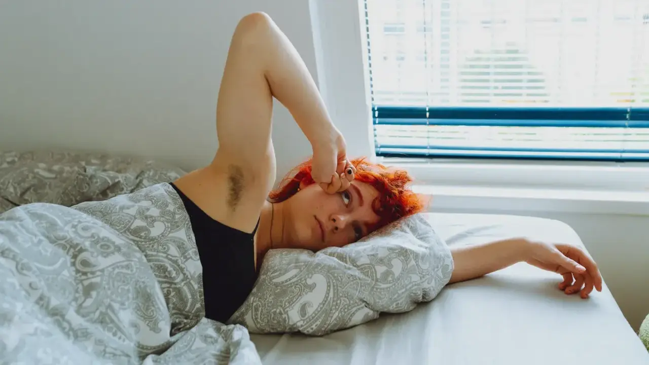 Jeune femme aux cheveux roux allongée dans son lit, le bras levé, le regard perdu. Elle semble fatiguée, peut-être un des symptômes manque magnésium.