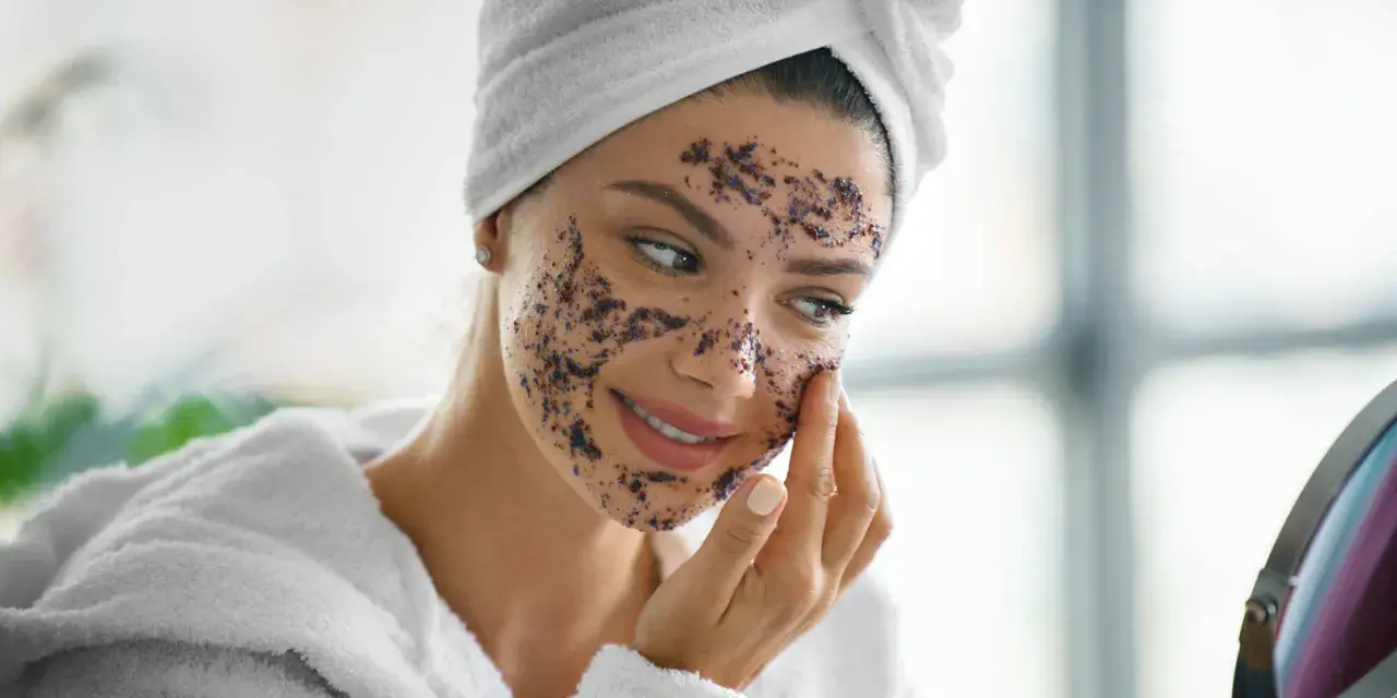 Une femme souriante applique un masque d'exfoliation visage. Elle porte une serviette sur la tête et un peignoir blanc.