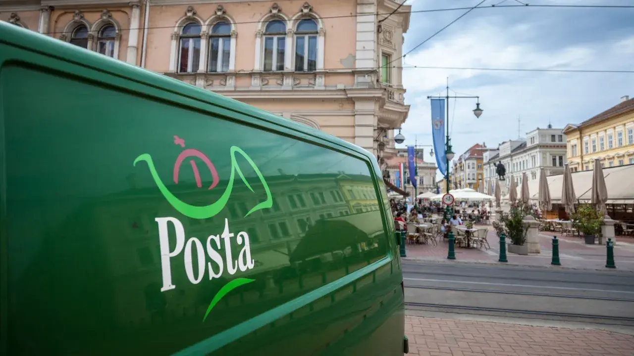 Zielona furgonetka pocztowa z logo "Posta" stoi na ulicy w węgierskim mieście. W tle widać kawiarnie i zabytkowe budynki.